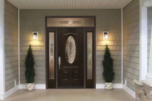 Flanked by two outside lights and plants, is a dark brown front entry door with an oval glass insert